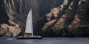 Sailboat gliding by the dramatic Na Pali Coast cliffs in Hawaii's clear waters.