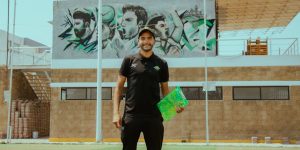 Smiling soccer coach stands on field with sports mural in the background, promoting training and sportsmanship.