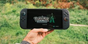 A hand holding a handheld gaming console displaying the Pokémon Legends game screen outdoors.